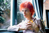 A color photograph of David Bowie with red hair, seated near a window, wearing a striped shirt.