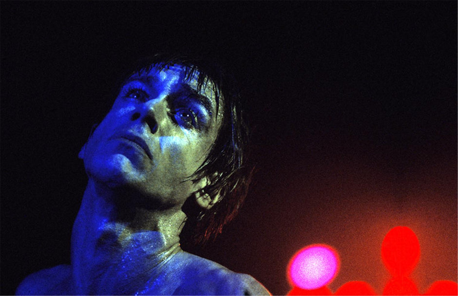 A fine art photo of Iggy Pop performing with a blue light on his face and blurred red and pink background lights. Photo shot by Mick Rock. 