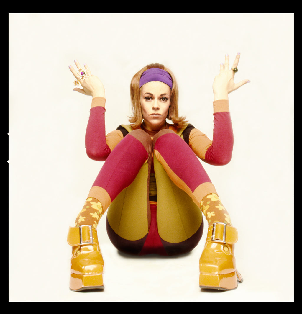 A photograph of Lady Miss Kier of Deee-Lite posing sitting with legs bent, wearing colorful tights and a yellow boots, with a purple headband.