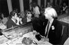 A black and white photograph of Mick Jagger and Andy Warhol seated at a dinner party, with Jagger smiling and Warhol wearing sunglasses.
