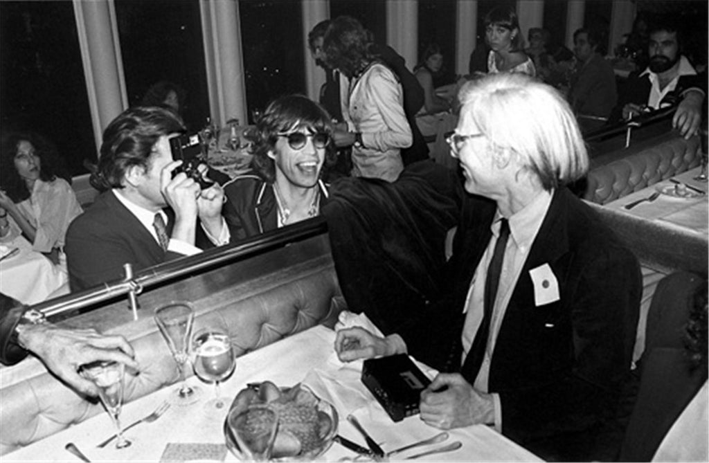 A black and white photograph of Mick Jagger and Andy Warhol seated at a dinner party, with Jagger smiling and Warhol wearing sunglasses.