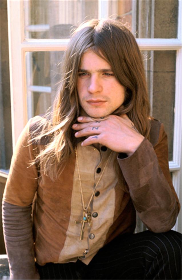 A black and white photograph of young Ozzy Osbourne, with his hand touching his chin, wearing a brown jacket and sitting by a window. shot by mick rock, 