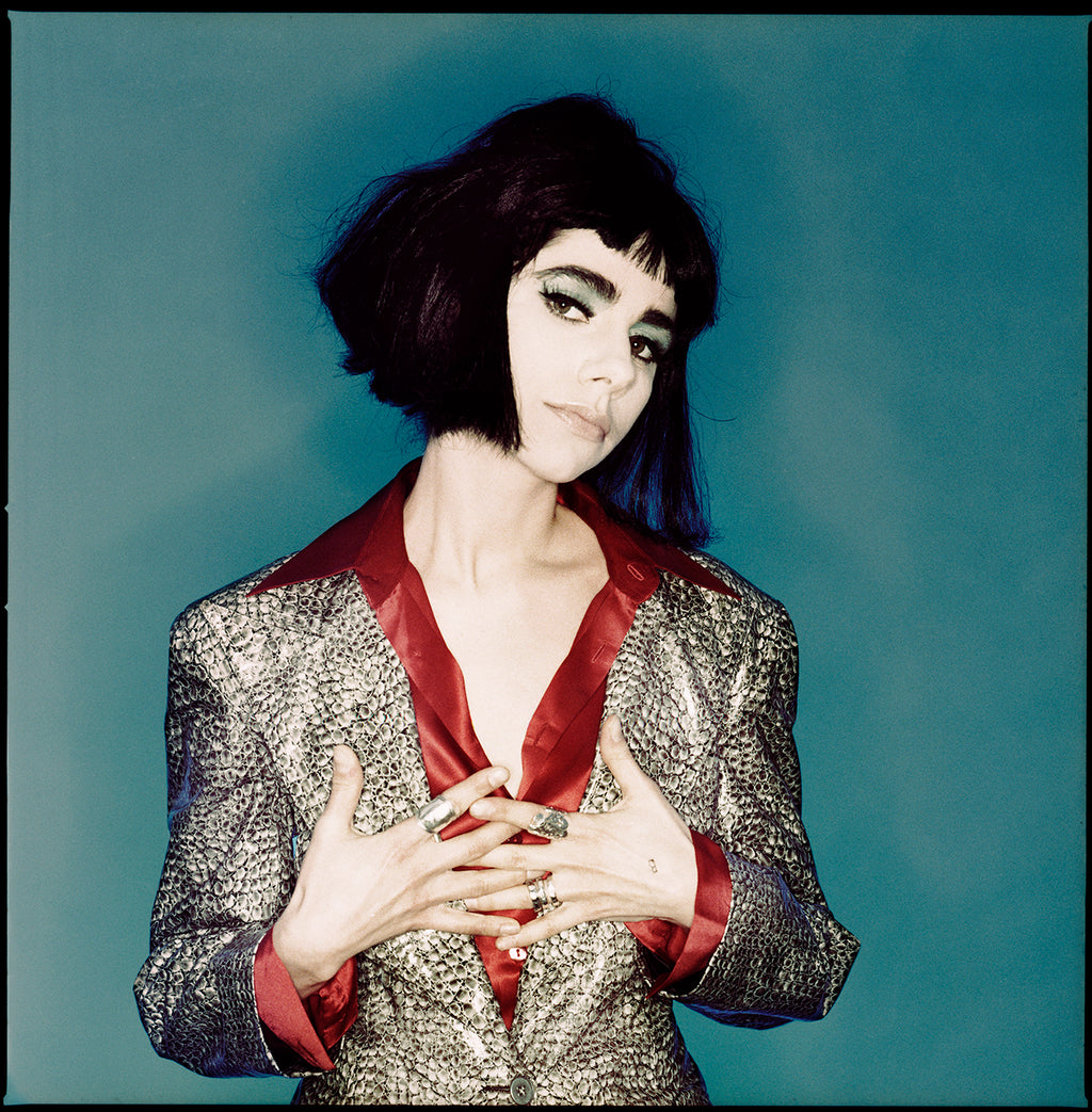 Fine art photo of singer PJ Harvey with cropped black hair, green eyeshadow, hands folded to her chest with red silk top and blue background. 