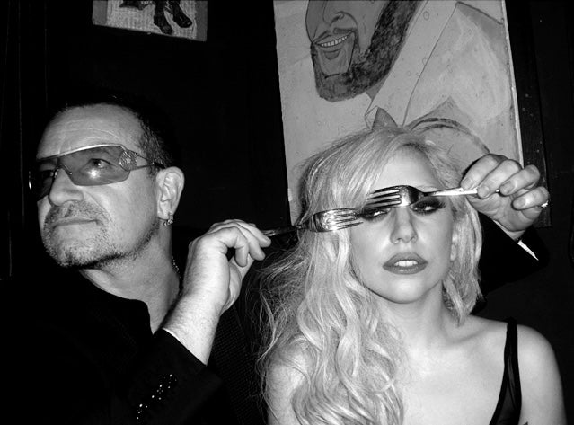 a black and white photo of bono of U2 and lady gaga. he is holding forks over her eyes. shot by photographer Mick Rock. 