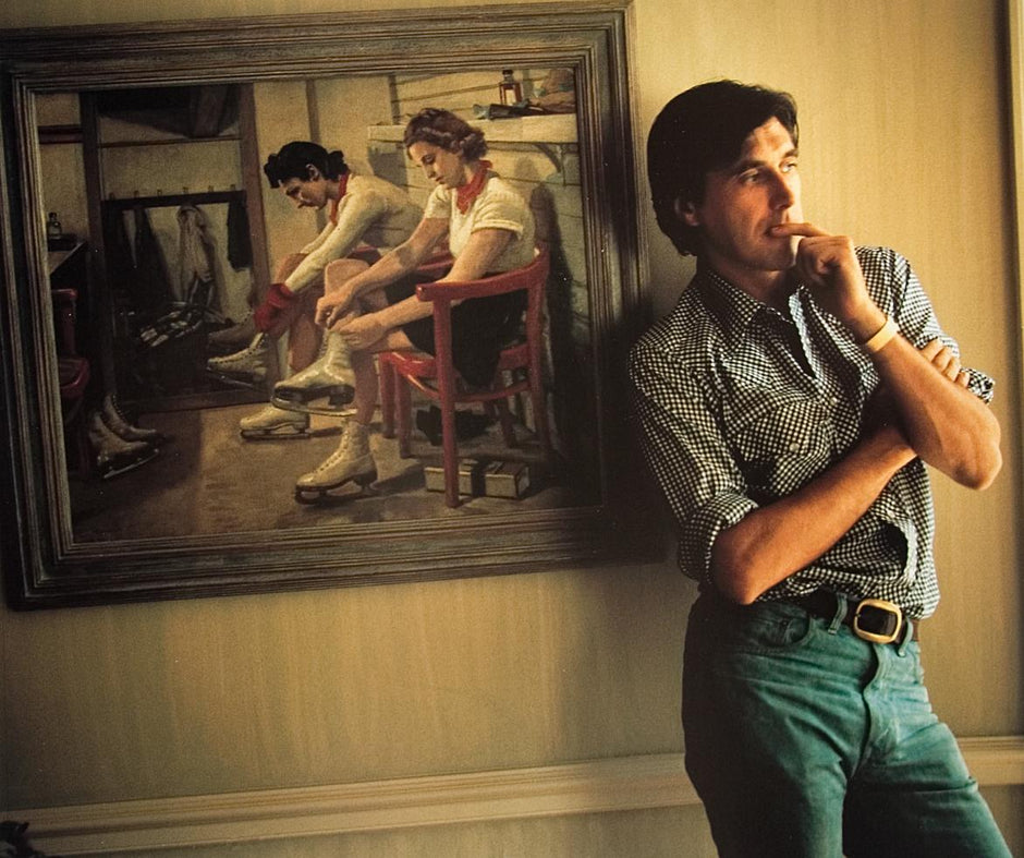 A photograph of Bryan Ferry of Roxy Music with a contemplative expression, seated in front of a painting, wearing a striped shirt and jeans. Photo by Mick Rock. 