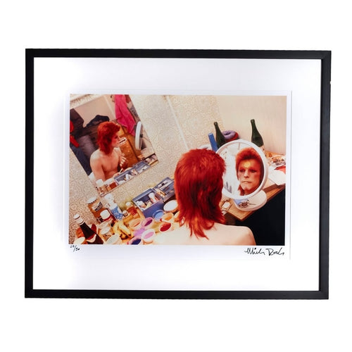 A fine art framed print of david bowie putting on makeup with bright red hair  in a circle mirror. Signed by the photographer Mick Rock. 