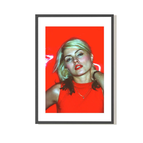 A photograph of Debbie Harry in a red dress, with hand behind her neck, against a red background. The image is a vintage Allan Tannenbaum photograph.