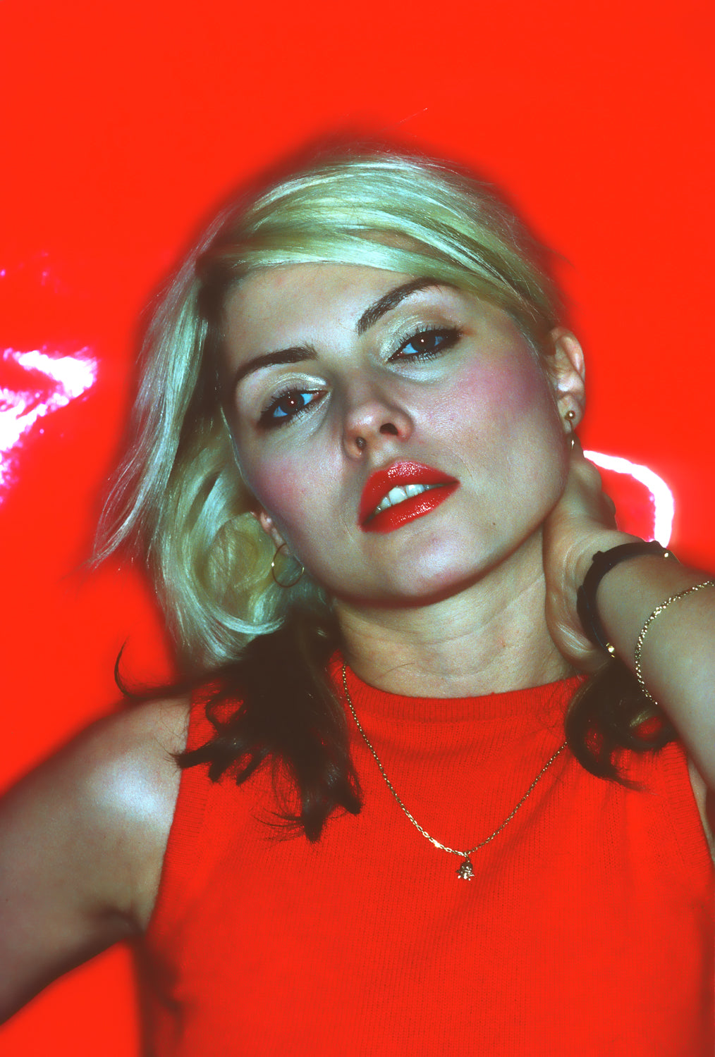 A photograph of Debbie Harry in a red dress, with hand behind her neck, against a red background. The image is a vintage Allan Tannenbaum photograph.