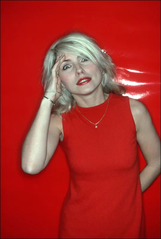 A photograph of Debbie Harry in a red dress, with a red salute, against a red background. The image is a vintage Allan Tannenbaum photograph.