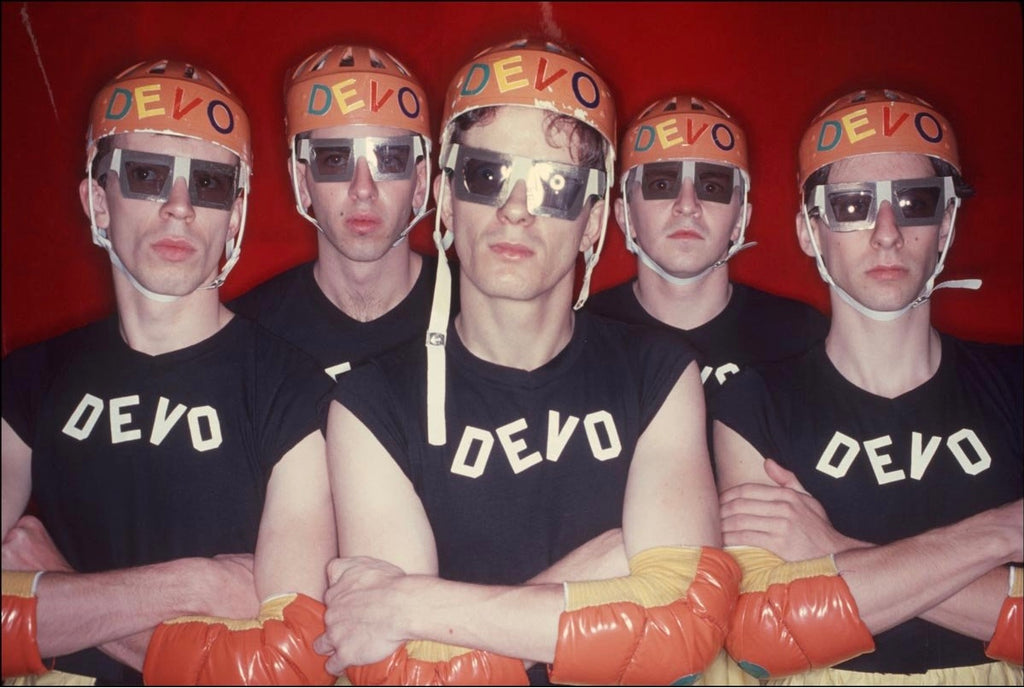 A group photograph of the Devo band members wearing similar black and orange outfits, posing with their iconic orange helmets.