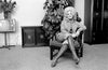 A fine art photo of Dolly Parton sitting in a chair living room setting with boots on, a plant and tv in the background. 