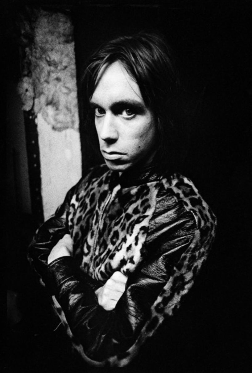 black and white portrait of Iggy Pop wearing a leopard print jacket with his arms crossed. Shot by photographer Mick Rock. 