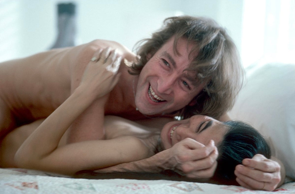 A fine art photo of John Lennon and Yoko Ono in bed, smiling at each other, with their arms around each other.