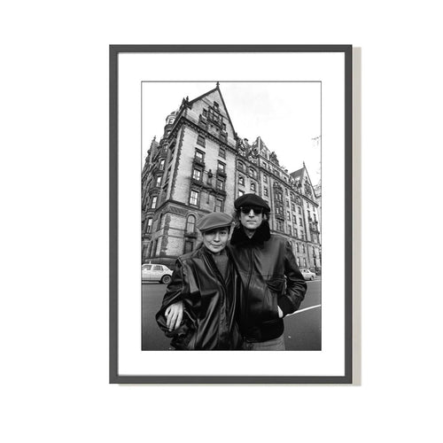 Black and white photograph of John Lennon and Yoko Ono outside the Dakota apartment house, posing with a building in the background.