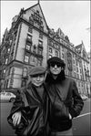 Black and white photograph of John Lennon and Yoko Ono outside the Dakota apartment house, posing with a building in the background.
