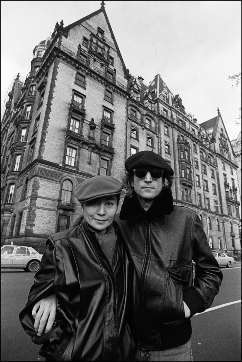 Black and white photograph of John Lennon and Yoko Ono outside the Dakota apartment house, posing with a building in the background.