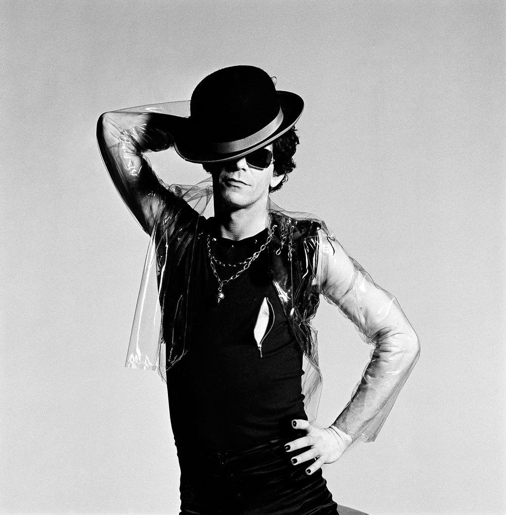 A black and white photograph of Lou Reed, taken by Mick Rock, with Reed posing in a theatrical manner, covering his face partially with a hand.