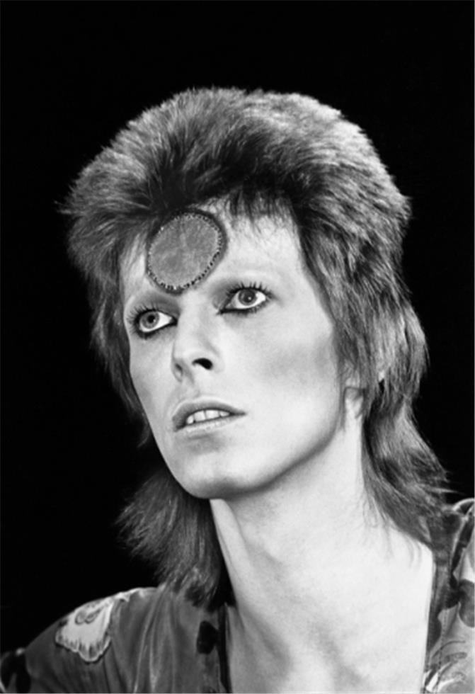 A black and white photograph of David Bowie, aka Ziggy Stardust, with a distinctive facial expression and makeup, wearing a glossy, dark outfit.