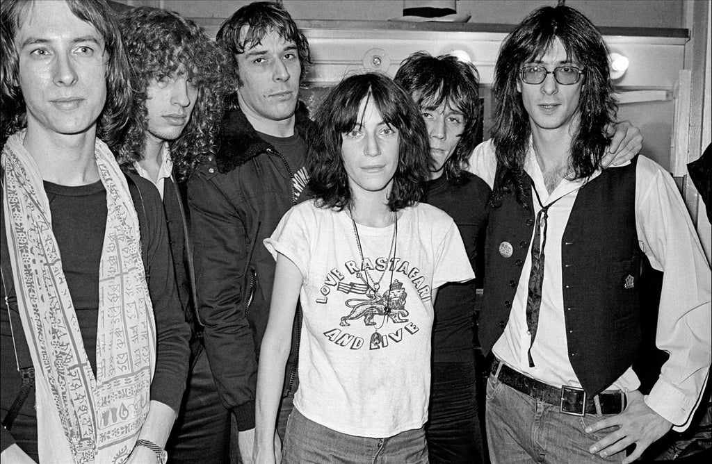 A black and white photograph of Patti Smith with John Cale, featuring them both in the foreground with other band members in the background.