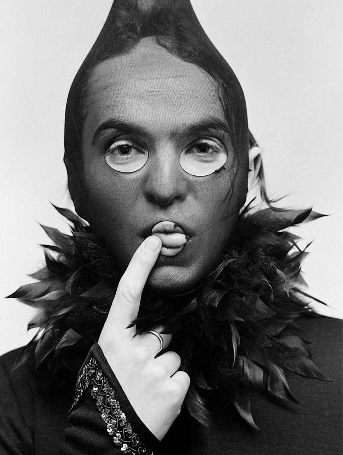 A black and white photograph of a person wearing black stocking and black feather boa around the neck. Shot by Mick Rock. 
