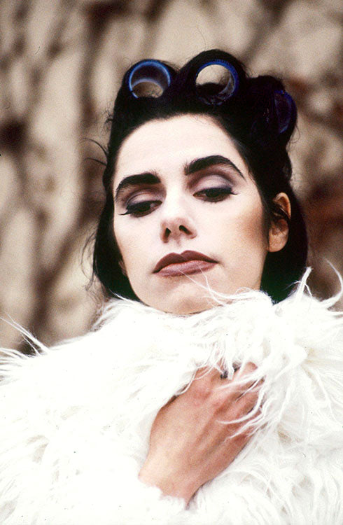 Photograph of PJ Harvey wearing white feathers, curlers in hair and dark eye makeup. 