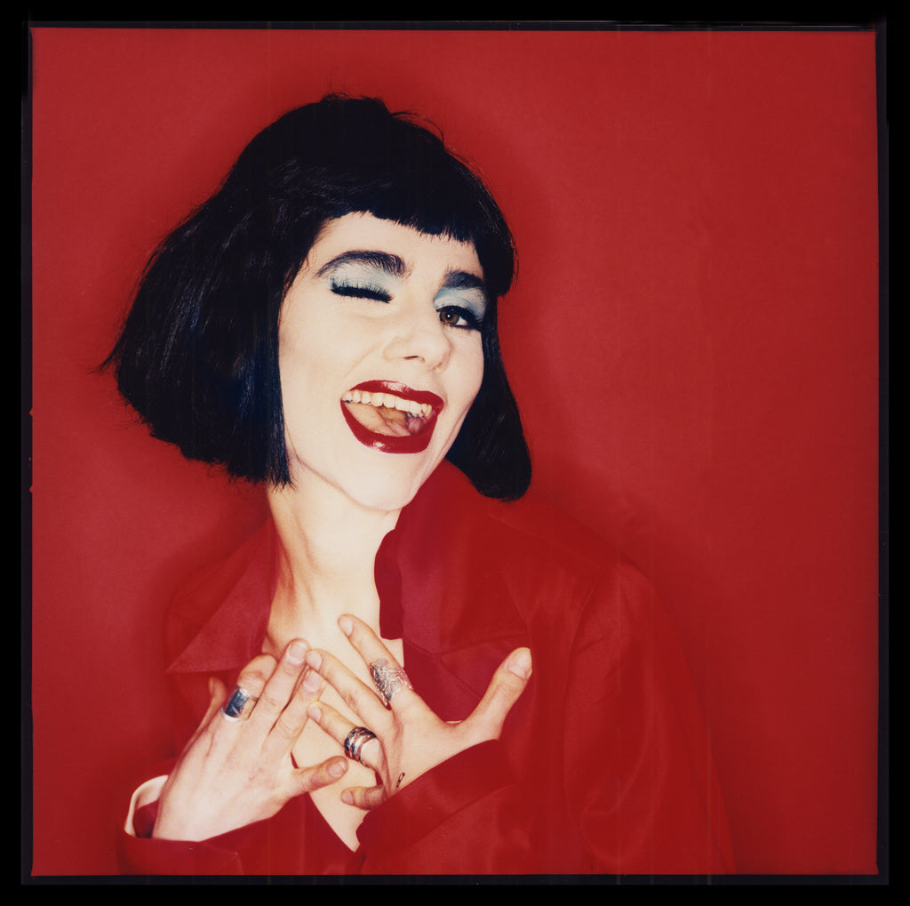 PJ Harvey shot winking with red background and black hair in London by photographer, Simon Fowler. This image was taken in Dorset at Poly’s Studio in 1995 for Select Magazine Cover. 