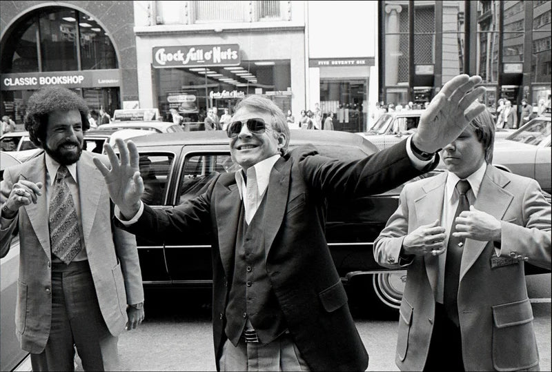 Black and white photograph of Steve Martin in a suit on a city street, extending his hand with a smile on his face, surrounded by buildings.