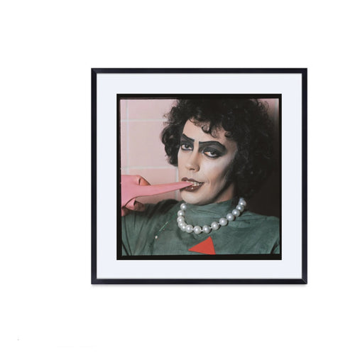A framed fine art photograph of Tim Curry, in a theatrical pose with a pink object in their mouth, makeup emphasizing their facial features, wearing a green outfit with a pearl necklace.