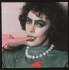 A fine art photograph of Tim Curry, in a theatrical pose with a pink object in their mouth, makeup emphasizing their facial features, wearing a green outfit with a pearl necklace.