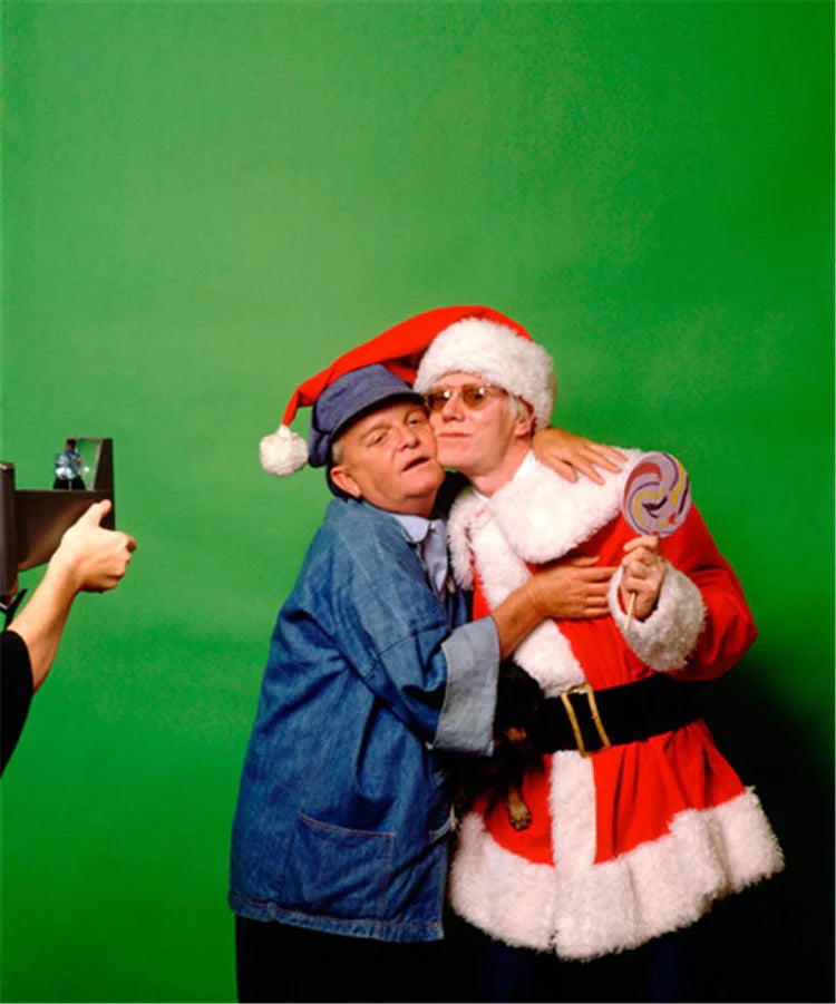 A photograph of Truman Capote and Andy Warhol wearing a Santa Claus hat hugging against a green background. 