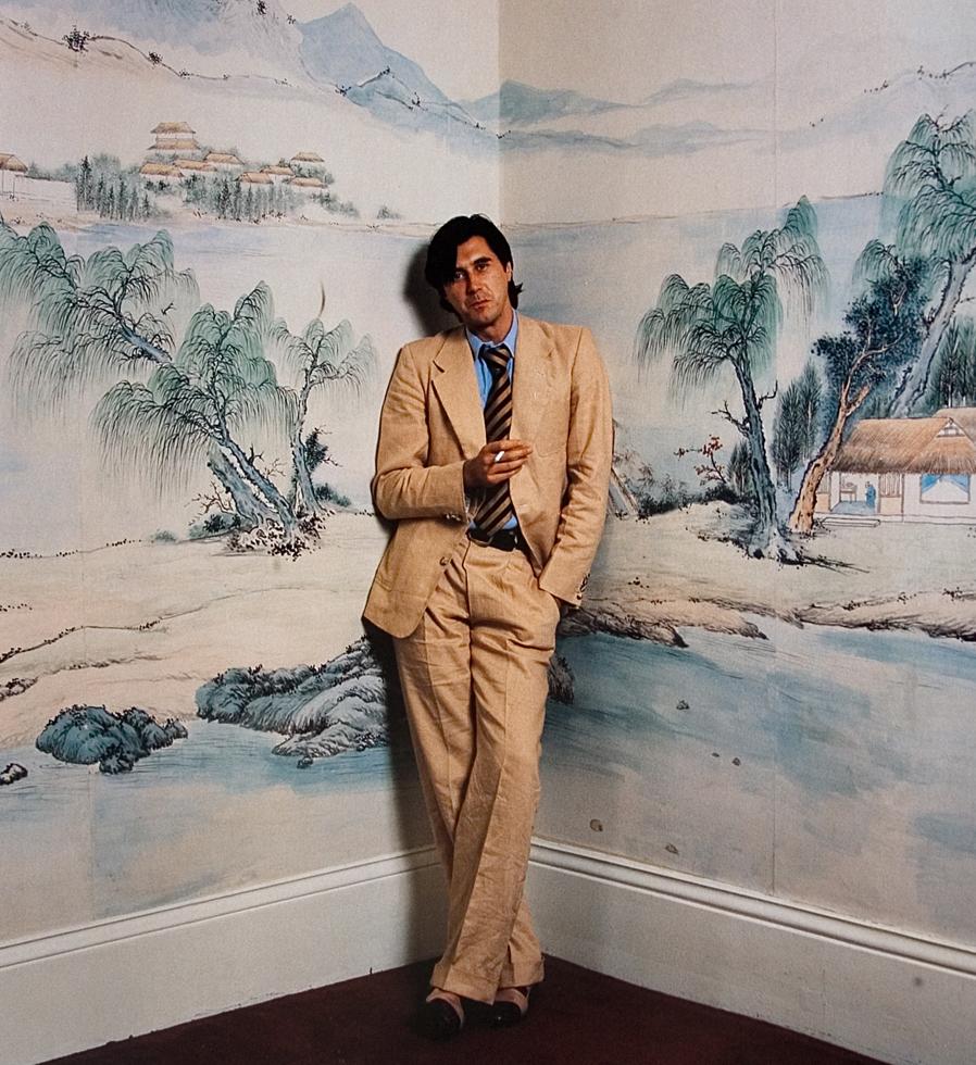 A fine art photograph of Bryan Ferry, in a contemplative pose with a blurred background featuring a landscape or architectural detail. Shot by Mick Rock. 